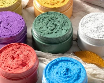 May include: A collection of round plastic jars filled with thick, textured cosmetic paste in various colours including yellow, green, purple, red, blue, and white. Each jar features a wide opening revealing the swirled, dense consistency of the product.
