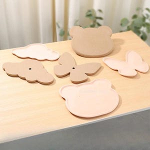 Puede incluir: Colección de bandejas decorativas con formas de nube, mariposas y cabezas de oso. Las bandejas son en tonos beige y rosa, dispuestas sobre una superficie de madera clara. Medidas en centímetros.