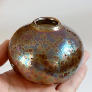 Puede incluir: Un pequeño jarrón redondo de cerámica con un esmalte metálico iridiscente. El jarrón tiene un patrón moteado en tonos marrones, dorados y azules. La abertura es de un color oscuro y reflectante. El jarrón se sostiene en una mano.