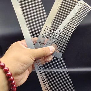 May include: Four metal stencils with a grid of small holes are displayed. Each stencil is rectangular with rounded corners and the number "139" printed on it. The stencils are held in a hand, and a red beaded bracelet is visible.