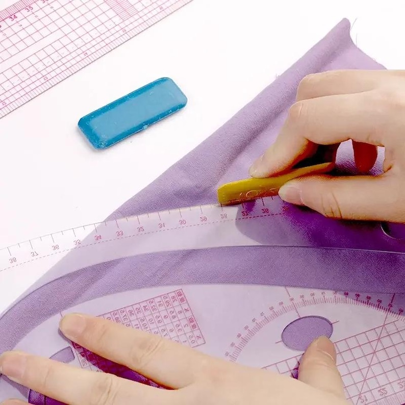 May include: A close-up of sewing tools and fabric. A clear, curved ruler is positioned over purple fabric, with a yellow marking tool in use. A blue rectangular chalk is also present, along with a pink and white grid ruler.
