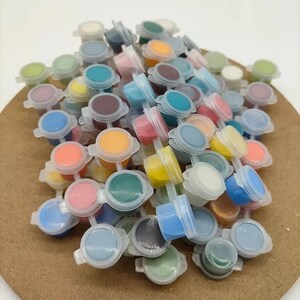 K&ouml;nnte beinhalten: Eine Sammlung kleiner Plastikfarbt&ouml;pfe, gef&uuml;llt mit verschiedenen Farben, darunter Blau, Rosa, Gelb und Gr&uuml;n. Die T&ouml;pfe sind auf einer braunen Oberfl&auml;che angeordnet, mit ge&ouml;ffneten Deckeln, die die Farbe zeigen. Das Bild zeigt eine Vielzahl von Farben.
