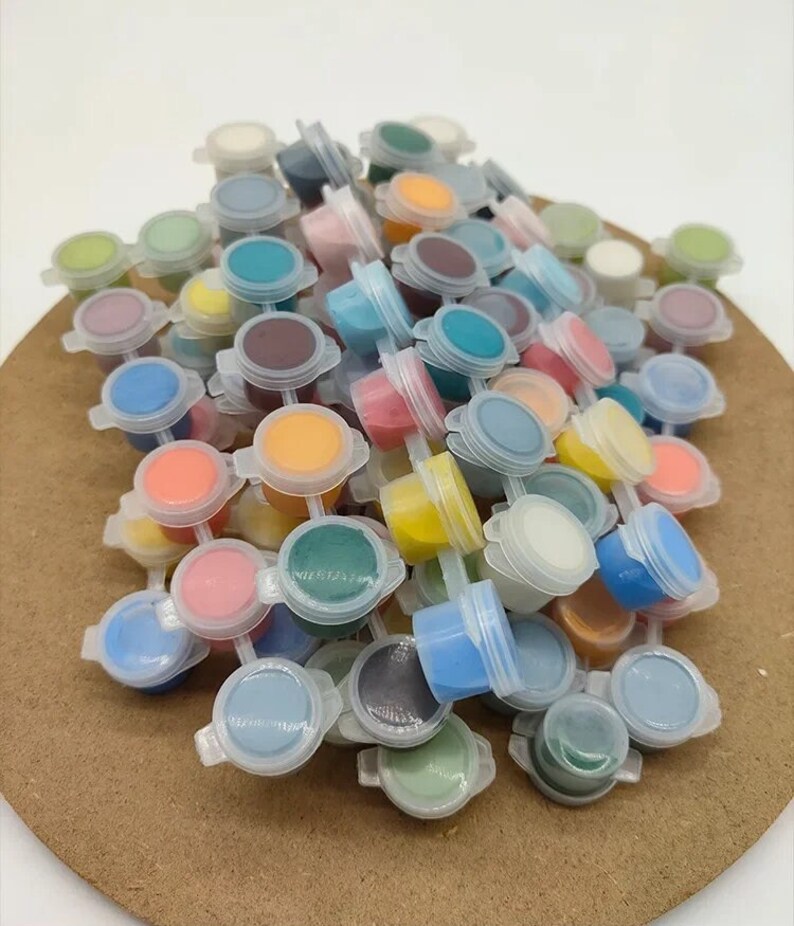 K&ouml;nnte beinhalten: Eine Sammlung kleiner Plastikfarbt&ouml;pfe, gef&uuml;llt mit verschiedenen Farben, darunter Blau, Rosa, Gelb und Gr&uuml;n. Die T&ouml;pfe sind auf einer braunen Oberfl&auml;che angeordnet, mit ge&ouml;ffneten Deckeln, die die Farbe zeigen. Das Bild zeigt eine Vielzahl von Farben.