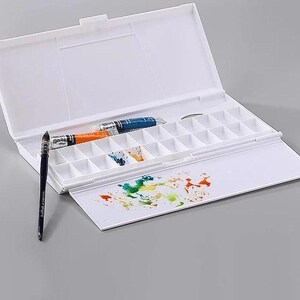 Op de afbeelding: Een witte, open aquarelverfpalet met een scharnierend deksel. Het palet bevat meerdere kleine compartimenten, twee tubes verf, een penseel en een mengruimte met kleurrijke verfspatten. Het palet staat op een grijze ondergrond.
