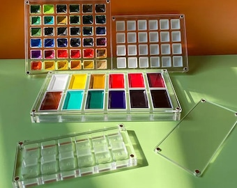 May include: Clear acrylic watercolor palettes filled with vibrant colors. The palettes are square and rectangular, with multiple compartments for paints. The colors include shades of red, yellow, blue, green, and purple. The palettes are displayed on a green surface.