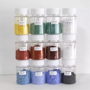 Könnte beinhalten: Eine Sammlung kleiner, klarer Gläser mit weißen Deckeln, die jeweils mit einem unterschiedlich farbigen Pulver gefüllt sind. Zu den Farben gehören Gelb, Grün, Blau, Rot und Weiß. Jedes Glas hat ein kleines weißes Etikett mit schwarzem Text.