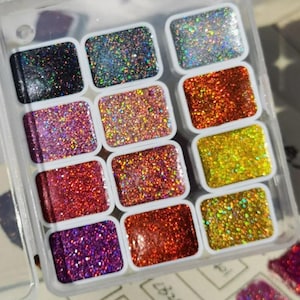 May include: A selection of twelve glitter eyeshadows presented in a clear plastic case. The eyeshadows are rectangular and feature a range of colours, such as black, blue, pink, orange, red, and yellow. Each eyeshadow is filled with glitter.