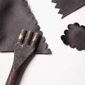 May include: A close-up of a metal leather punch tool with three prongs. The tool is lying on top of two pieces of black leather.