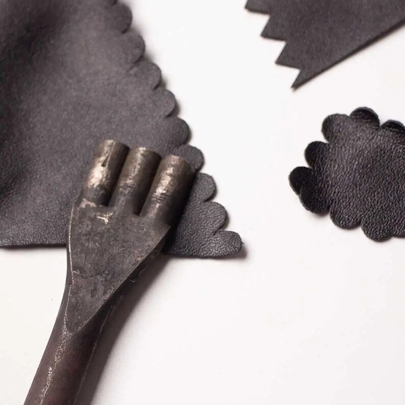 May include: A close-up of a metal leather punch tool with three prongs. The tool is lying on top of two pieces of black leather.