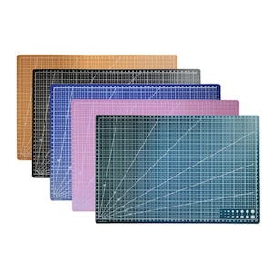 Op de afbeelding: Een stapel snijmatten in verschillende kleuren: bruin, zwart, blauw, roze en teal. Elke mat heeft een rasterpatroon met centimeter-metingen en hoeklijnen. Deze matten zijn ontworpen voor knutsel- en naaiprojecten.