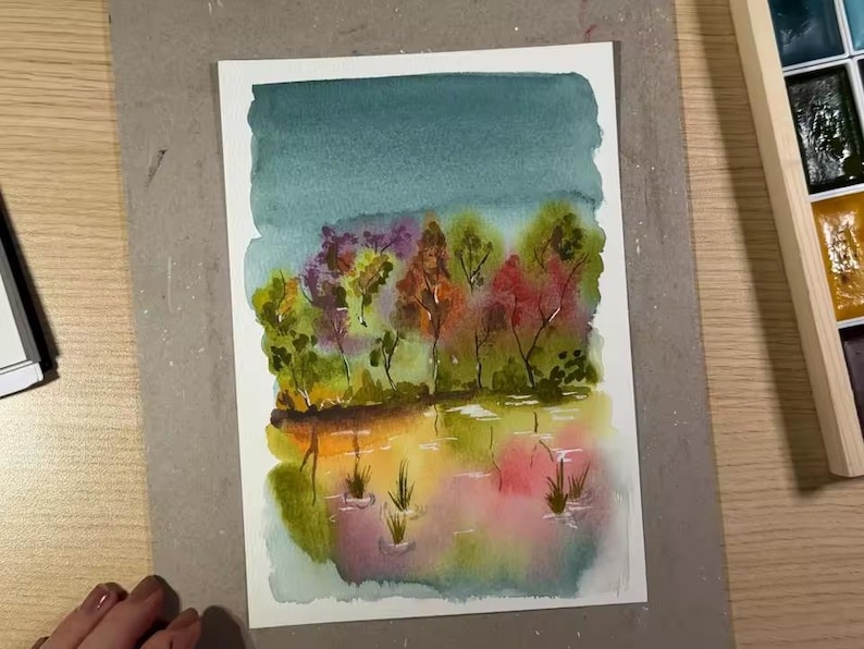 May include: Watercolor painting of a landscape with a teal sky, colorful trees, and a reflective body of water. The trees feature shades of green, red, and orange. The water reflects the colors of the trees and sky, with hints of pink and yellow.