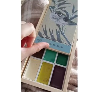 May include: A wooden watercolor paint set with a hinged lid featuring a bird illustration. The set contains various colors, including green, yellow, and brown. The lid is being opened, revealing the paint colors inside.