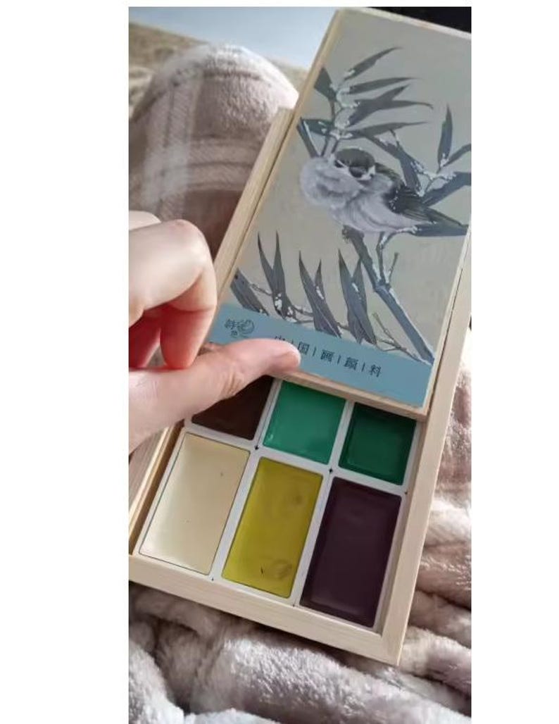 May include: A wooden watercolor paint set with a hinged lid featuring a bird illustration. The set contains various colors, including green, yellow, and brown. The lid is being opened, revealing the paint colors inside.