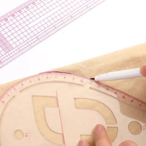 May include: A clear plastic pattern-making ruler and a pink ruler are positioned over tan fabric. A white pen is used to trace a curved line. The rulers have measurements in inches and are used for sewing and design.