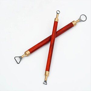 May include: Two red-brown wooden clay sculpting tools with brass accents and black metal loops and blades. The tools are crossed against a white background. The tools are used for pottery and sculpting.