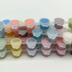 K&ouml;nnte beinhalten: Eine Sammlung kleiner, klarer Plastikfarbt&ouml;pfe, gef&uuml;llt mit verschiedenen Farben wie Blau, Rosa, Gelb und Gr&uuml;n. Die T&ouml;pfe sind in einem transparenten Kunststoff-Organizer gestapelt, was eine ansprechende Pr&auml;sentation von Kunstbedarf ergibt.