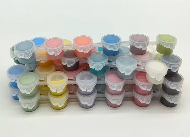 K&ouml;nnte beinhalten: Eine Sammlung kleiner, klarer Plastikfarbt&ouml;pfe, gef&uuml;llt mit verschiedenen Farben wie Blau, Rosa, Gelb und Gr&uuml;n. Die T&ouml;pfe sind in einem transparenten Kunststoff-Organizer gestapelt, was eine ansprechende Pr&auml;sentation von Kunstbedarf ergibt.