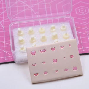 May include: A clear plastic container holding several small, off-white dental molds. A beige rectangular pad with pink lip-shaped designs is in front of the container. The background is a pink cutting mat.