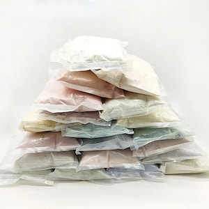 May include: A large pyramid-shaped stack of small, clear plastic pouches filled with various coloured powders. The collection includes shades of white, cream, pale pink, soft blue, light green, and muted grey, all neatly arranged in a tiered formation.