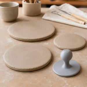 May include: Three flat, circular, light brown clay slabs of varying sizes arranged on a surface, accompanied by a light grey circular clay stamp with a spherical handle, a small clay cup, a cylindrical clay container, and two metal sculpting tools.