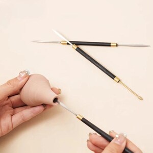 Peut inclure: Un récipient en argile brun clair de forme bulbeuse, sculpté à l'aide d'un outil en métal. Deux autres outils de sculpture à double extrémité, dotés de manches noirs et de viroles dorées, présentent des pointes métalliques fines et arrondies.