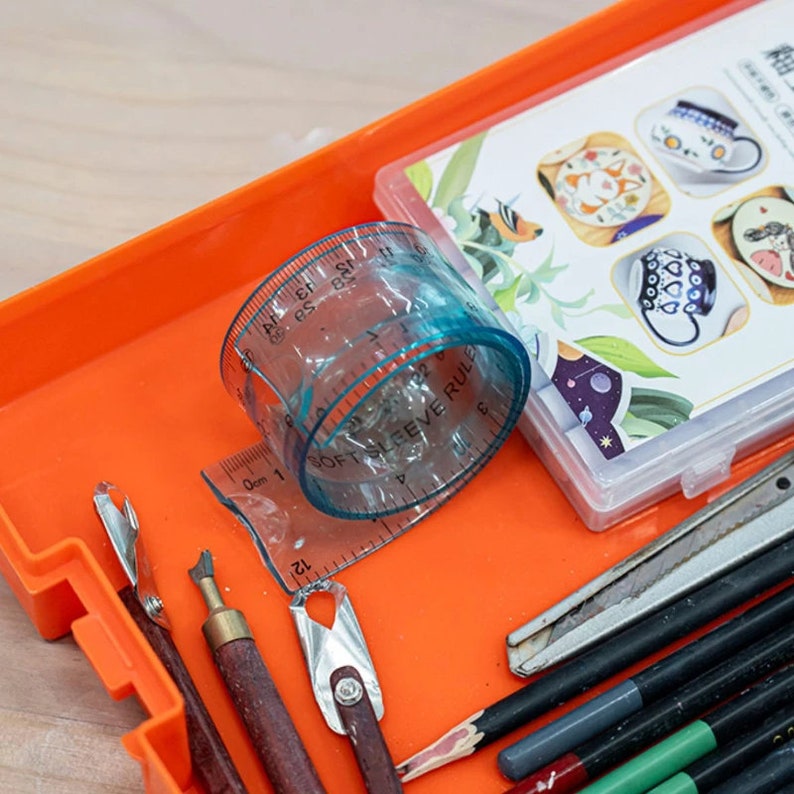 May include: An orange plastic tray with various art supplies, including a clear plastic ruler, pencils, and a small box with illustrations of mugs. The ruler is labeled "Soft Sleeve Rule" and has markings in centimeters and inches.