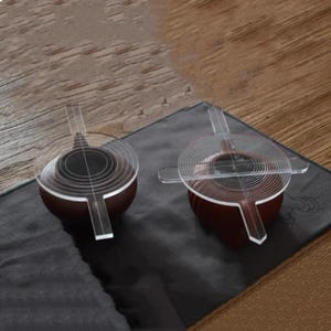 Könnte beinhalten: Zwei kleine, dunkelrote Teetassen mit transparenten, kreisförmigen Deckeln und Griffen werden auf einer dunkelgrauen Oberfläche präsentiert. Die Deckel haben konzentrische Kreise. Der Hintergrund ist ein Holztisch.