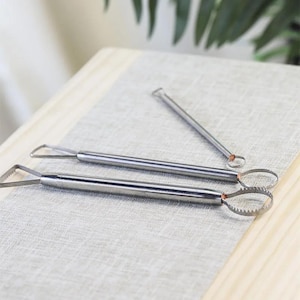 Pode incluir: Três ferramentas de escultura prateadas com pontas de diferentes formas. As ferramentas estão sobre uma superfície de tecido cinza claro.