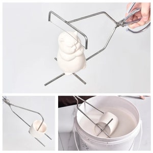 May include: A metal tool for dipping and holding ceramic objects. The tool has two arms that cross at the centre and a wire loop at the end of each arm. The tool is shown being used to dip a ceramic cat figurine into a bucket of white liquid.