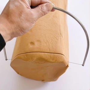Könnte beinhalten: Ein hellbrauner rechteckiger Tonblock wird mit einem Drahtschneider geschnitten. Der Schneider besteht aus Edelstahl und hat einen gebogenen Griff. Der Ton hat eine strukturierte Oberfläche und wird horizontal geschnitten.