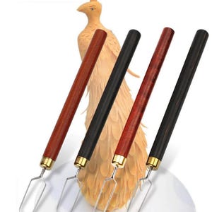 May include: Four clay sculpting tools with long cylindrical handles, two in reddish-brown wood and two in black wood, each featuring a gold-coloured metal ferrule and a silver-toned wire loop tip, arranged in front of a carved wooden peacock figure.