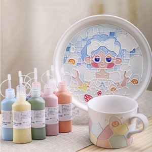 Puede incluir: Un juego de plato y taza de cerámica con un diseño caprichoso, que presenta a una niña de dibujos animados en una bañera. El plato tiene una combinación de colores azul y blanco, y la taza tiene un patrón geométrico. Seis botes de pintura de varios colores.