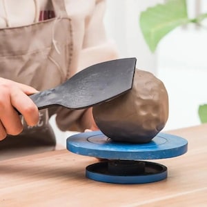 Puede incluir: Una persona utiliza una herramienta de plástico negro para dar forma a una bola de arcilla marrón en un torno de alfarería azul.