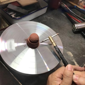 May include: A close-up of a pottery wheel with a small, brown clay sculpture in the center. A T-shaped tool is positioned near the clay, and a hand is holding the tool. The wheel is silver and has concentric circles.