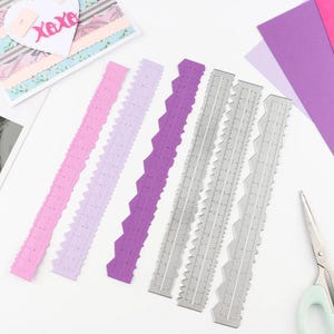 May include: A selection of decorative paper cutting rulers in shades of pink, lilac, purple, and silver. The silver rulers feature measurement markings. A greeting card with the text "XOXO" is visible in the background.