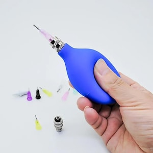Könnte beinhalten: Ein blauer Luftblasebalg aus Gummi mit einer Metalldüse und einem Satz bunter Nadeln. Der Blasebalg wird in einer Hand gehalten.