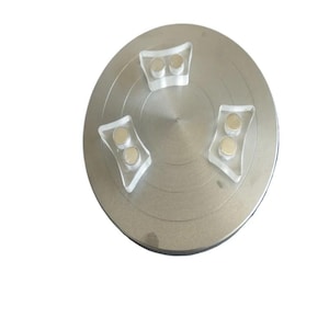 Może przedstawiać: Srebrny metalowy gramofon z trzema przezroczystymi akrylowymi wspornikami, każdy z dwoma małymi, okrągłymi magnesami. Gramofon ma koncentryczne rowki.