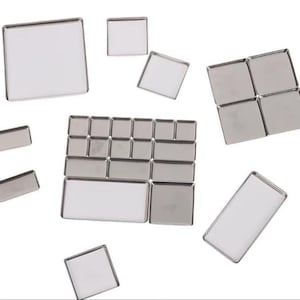 May include: A collection of various-sized, silver-toned metal trays with clear, recessed centers. The trays are rectangular and square, arranged against a white background. Some trays are grouped together, while others are scattered.