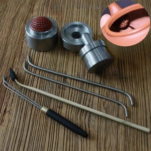 May include: A collection of tea ceremony tools, including a metal tea strainer with a red silicone insert, a metal tea press, and several stainless steel and wooden tea utensils. The tools are arranged on a wooden surface.
