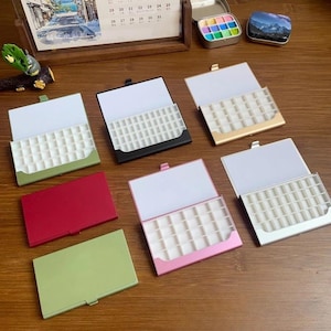 Op de afbeelding: Een verzameling open, rechthoekige metalen koffers in verschillende kleuren, waaronder groen, rood, roze, zwart en goud. Elke koffer heeft een wit interieur met kleine, vierkante compartimenten. Een kalender en kleine decoratieve blikjes zijn ook zichtbaar.