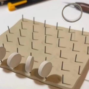 Könnte beinhalten: Ein rechteckiges beiges Brett mit einem Gitter aus aufrechten Metallstiften. Vier weiße kreisförmige Scheiben sind auf der vorderen Stiftreihe montiert. Eine kleine Spule aus dünnem Silberdraht befindet sich in der oberen rechten Ecke.