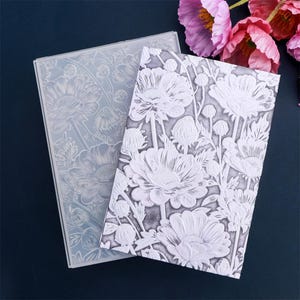 May include: Two rectangular floral-patterned craft stencils. One is clear, the other is white with a raised design. The floral design features large flowers and buds. The stencils are on a dark blue surface.