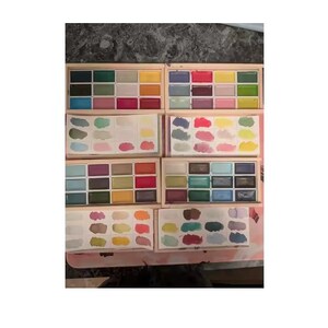May include: A collection of watercolor paint sets in wooden boxes. Each box contains an array of vibrant colors, including shades of green, blue, pink, red, and yellow. The sets are arranged in a grid-like pattern, showcasing the variety of hues.