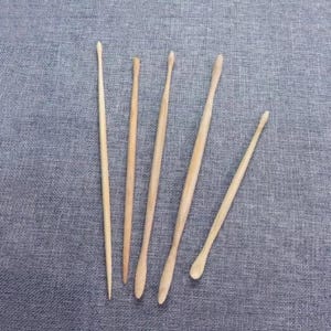 Puede incluir: Un conjunto de cinco baquetas de madera de diferentes longitudes y formas, dispuestas sobre una superficie de tela gris. Las baquetas son de color marrón claro, con puntas redondeadas o puntiagudas.