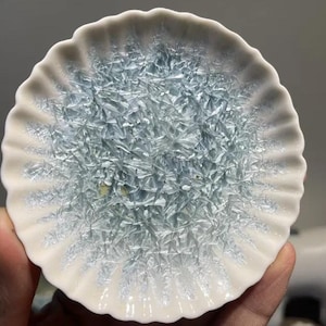 May include: A small, round, ceramic dish with a ruffled edge. The dish features a light blue, crystalline pattern in the center, surrounded by a white border. The dish is held by a hand.