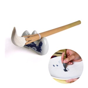 May include: A white brush with a bamboo handle resting on a white ceramic brush holder with blue and white floral designs. The brush is resting on the holder with the bristles facing up. A close-up of a hand using the brush to paint on a white plate with blue and white floral designs.