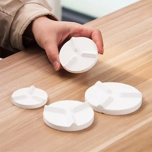 Puede incluir: Un conjunto de tres objetos circulares blancos con un diseño en relieve de tres puntas en la superficie superior. Los objetos varían en tamaño, uno se sostiene y dos descansan sobre una superficie. Cada pieza tiene un acabado mate y liso.