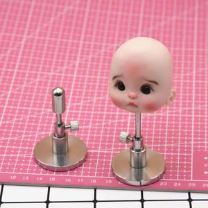 May include: Two doll head stands with metal bases. The stands are silver and have a screw to adjust the height. The doll heads are not included.