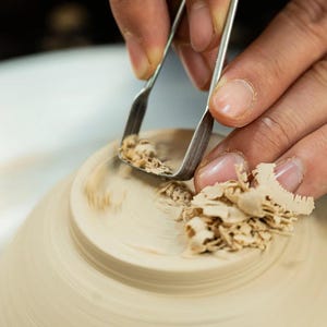 Pode incluir: Uma ferramenta de metal para cerâmica é usada para esculpir um anel de base circular no fundo de uma tigela de argila bege clara sem esmalte. Pequenas aparas finas de argila acumulam-se na superfície à medida que o material é removido.