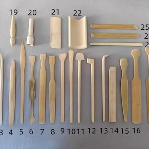 Peut inclure: Divers outils de cérémonie du thé en bambou, dont une louche, un fouet et diverses spatules et agitateurs. Les outils sont disposés sur une surface gris clair, mettant en valeur leurs tons de bois naturels et leurs formes variées. Collection numérotée de 1 à 25.